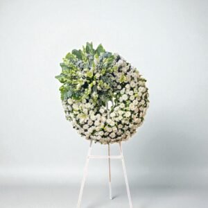 Corona fúnebre blanca con lirios, flor argentina y follaje verde sobre tripie de madera.