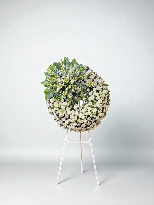 Corona fúnebre blanca con lirios, flor argentina y follaje verde sobre tripie de madera.